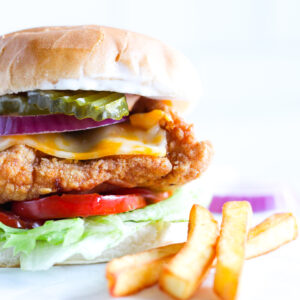 Fried chicken sandwich on a bun with cheese, lettuce, pickles, tomatoes and fries on the side.