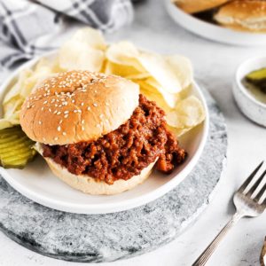 A plate containing a sloppy Joe on a bun, pickles, and chips.