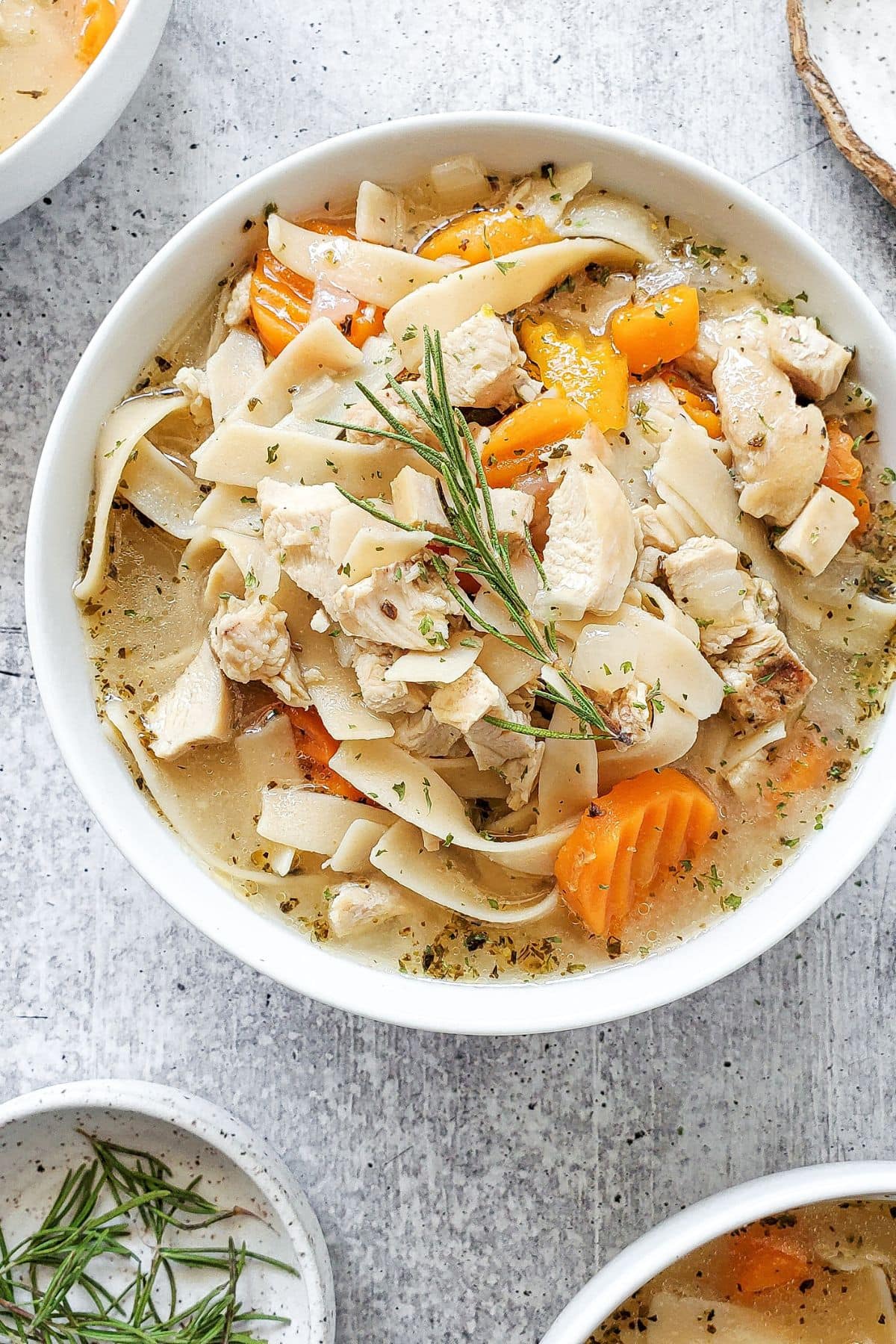 A white bowl with chicken noodle soup in a flavorful broth, garnishes with fresh rosemary herb.