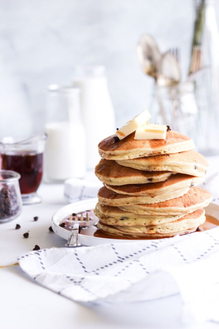 A fluffy stack of pancakes with 2 pats of butter on top on a white plate with syrup. Milk, syrup, and chocolate chips are also in the image to the left.