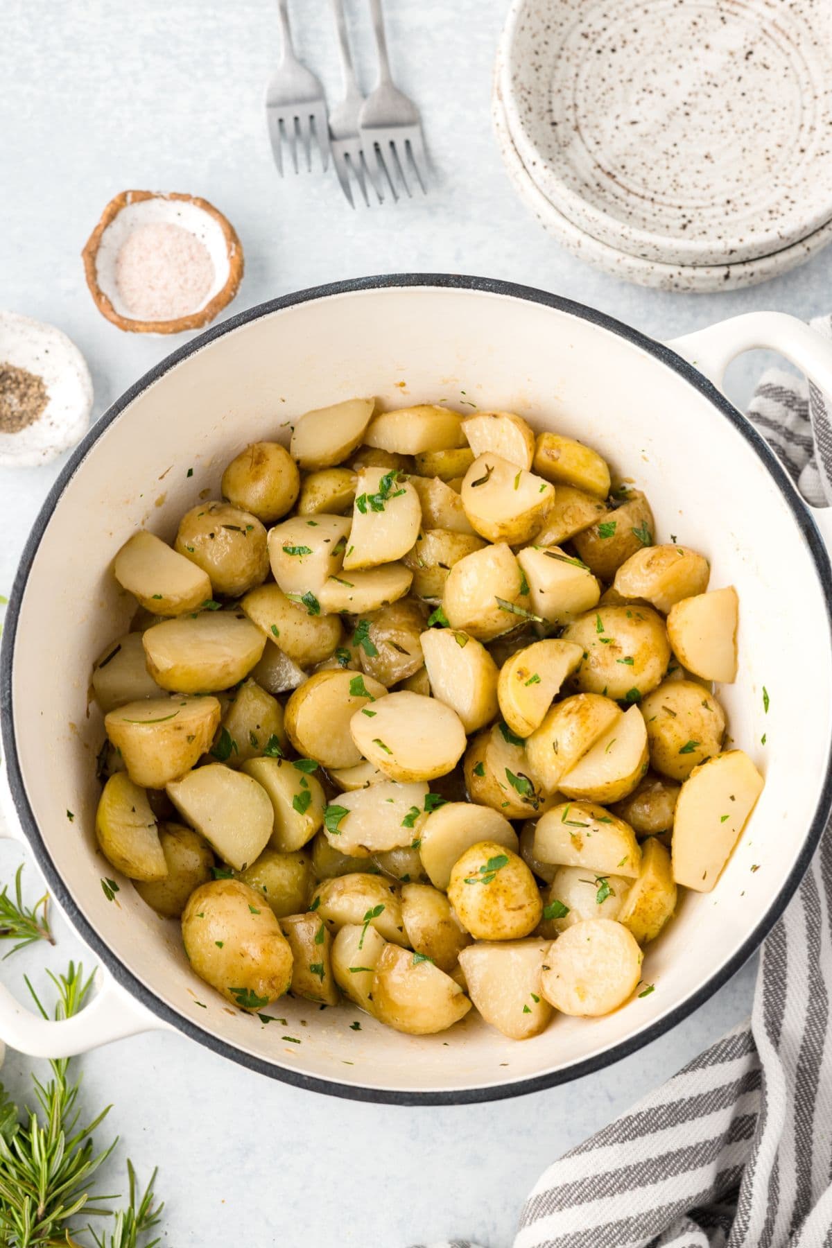 Sliced, herbed potatoes in a large white dutch oven surrounded by a linen and fresh herbs.