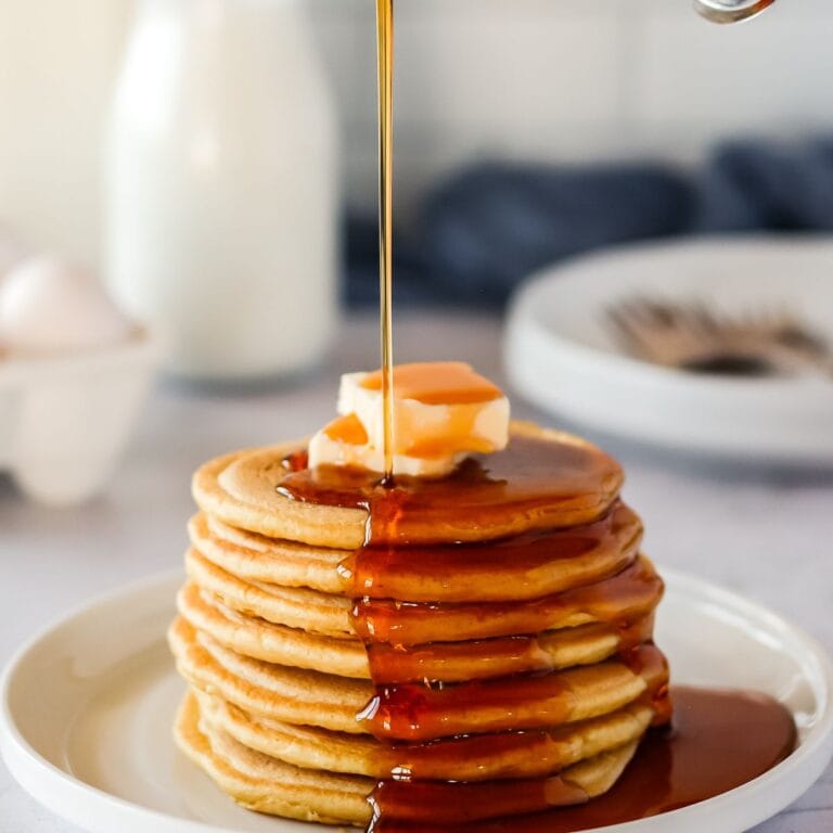 A fluffy stack of sourdough pancakes on a white plate with a drizzle of maple syrup being poured on them.