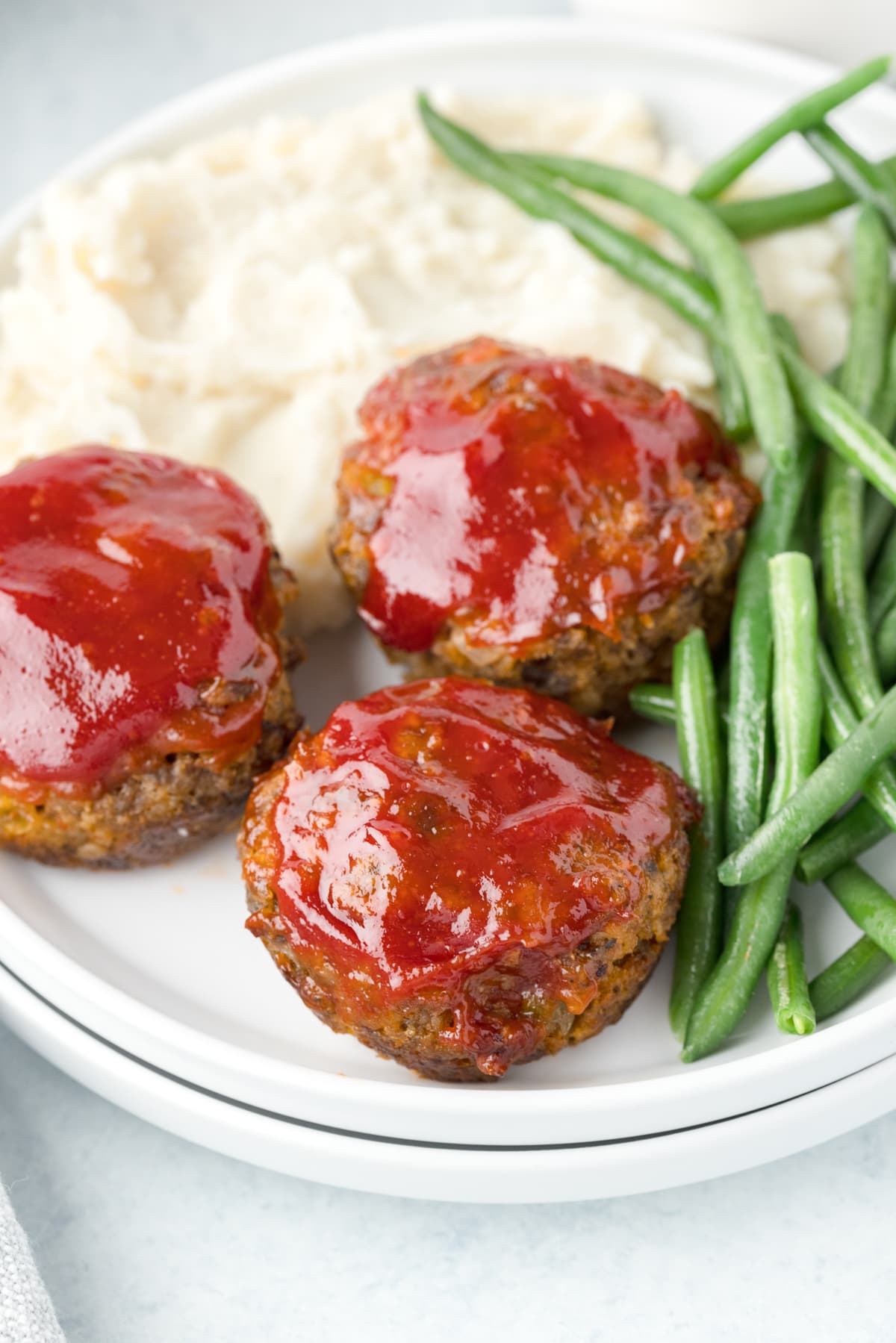 3 mini meatloaf muffins on a stacked white plae with long green beans and mashed potatoes