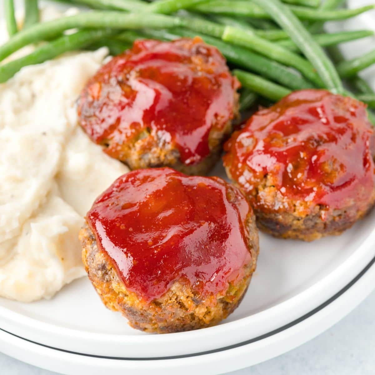 miniature meatloaves with a sweet ketchup and brown sugar glaze on white stacked plates served with mashed potatoes and green beans.