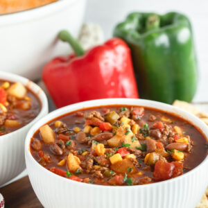 A white bowl filled with cowboy, and one is offset to the left with a large white dutch oven in the background with red and green bell peppers.