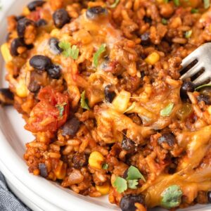 A close-up view of a Mexican Beef and Rice Skillet meal with melted cheese, black beans, corn, and fresh cilantro, with a fork digging in.