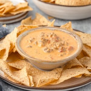 A bowl of creamy cheese dip with sausage and diced tomatoes in a small bowl on a large plate, accompanied by a plate of tortilla chips.