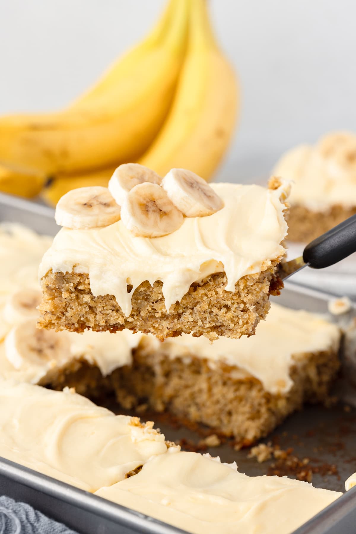 A spatula holds up a slice of banana cake with cream cheese frosting, topped with banana slices.