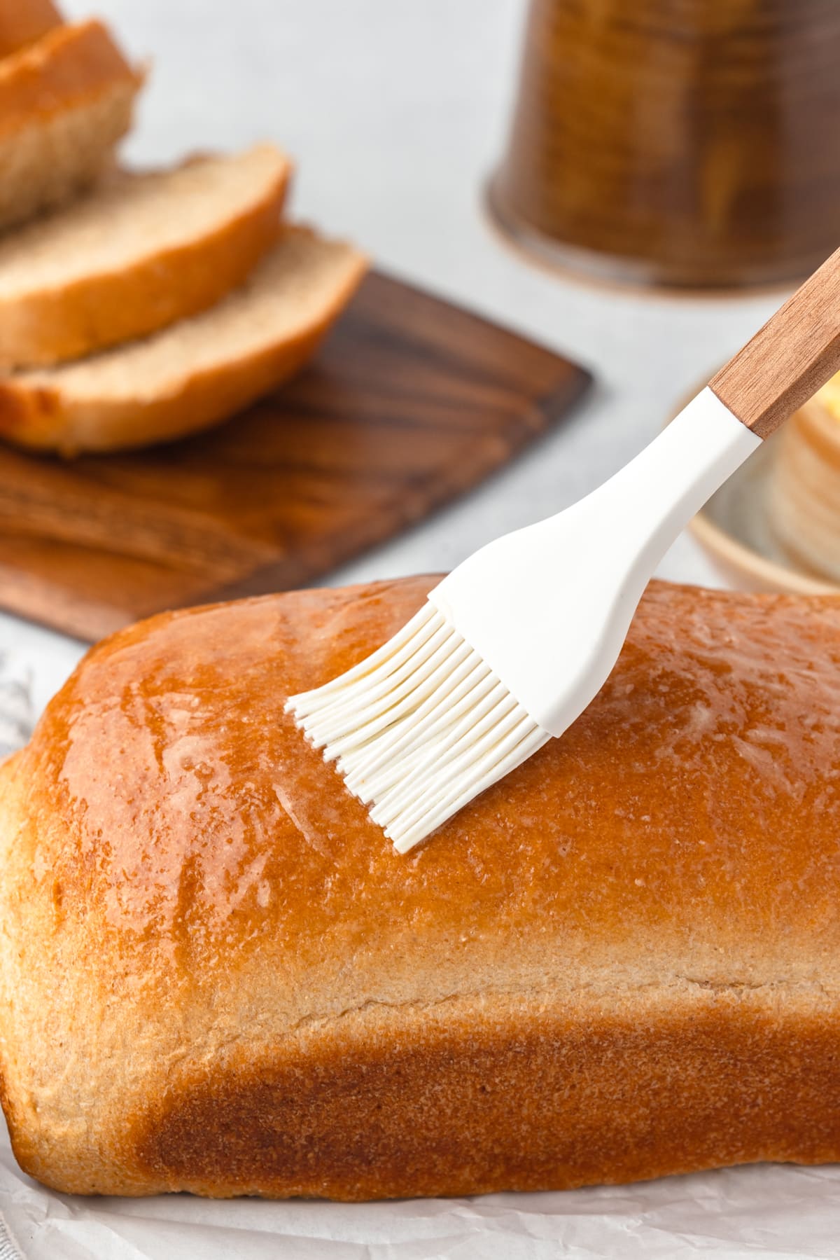 An uncut loaf of whole wheat bread getting brushed with melted butter with a white pastry brush.