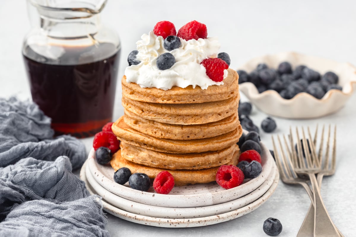 A stack of whole wheat pancakes on white stoneware plates topped with whipped cream and fresh fruit.