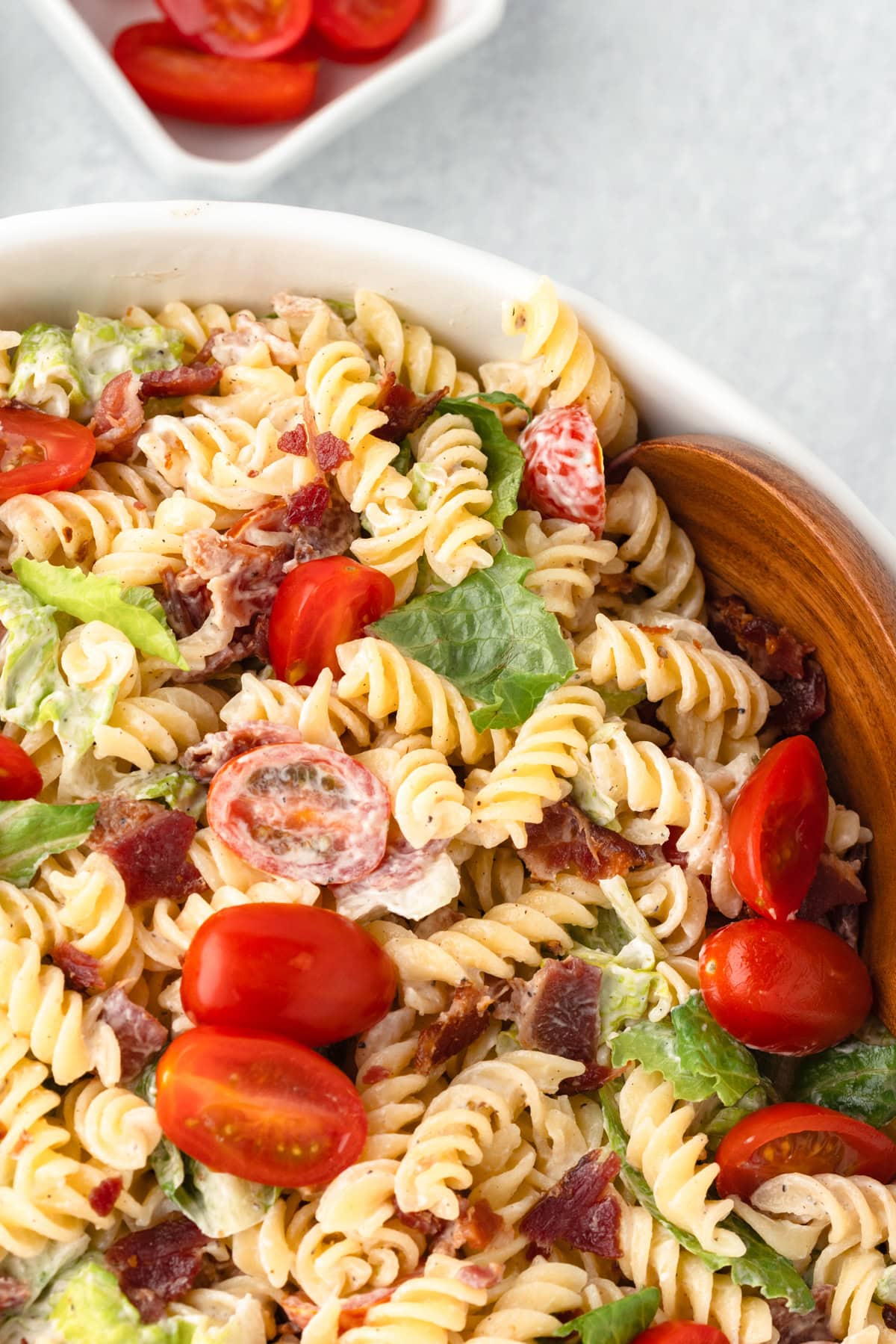 A large white bowl containing a blt pasta salad with large wooden spoons. It has bacon, romaine lettuce and grape tomatoes.