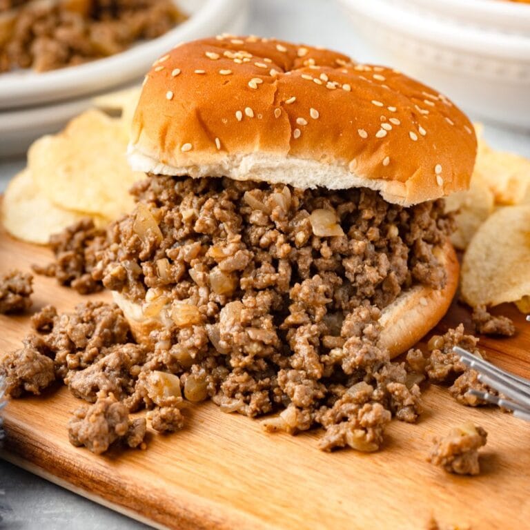 Loose meat sandwich on a wooden board. The beef mixture is spilling out of the hamburger bun, with chips in the back.