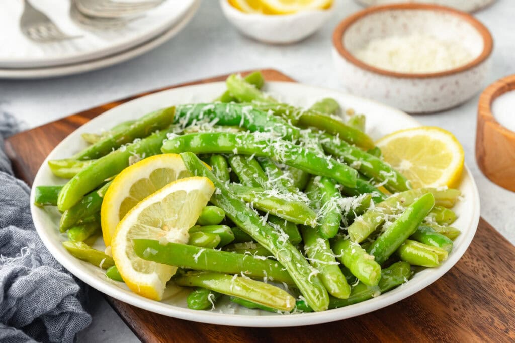A plate of fresh green beans garnished with grated cheese and lemon slices. The dish is placed on a wooden board, with small bowls of seasoning and a stack of plates visible in the background.