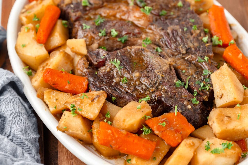A plate of roast beef surrounded by chunks of roasted potatoes, carrots, and garnished with chopped parsley. The dish is presented on a white oval plate with a rustic wooden background.