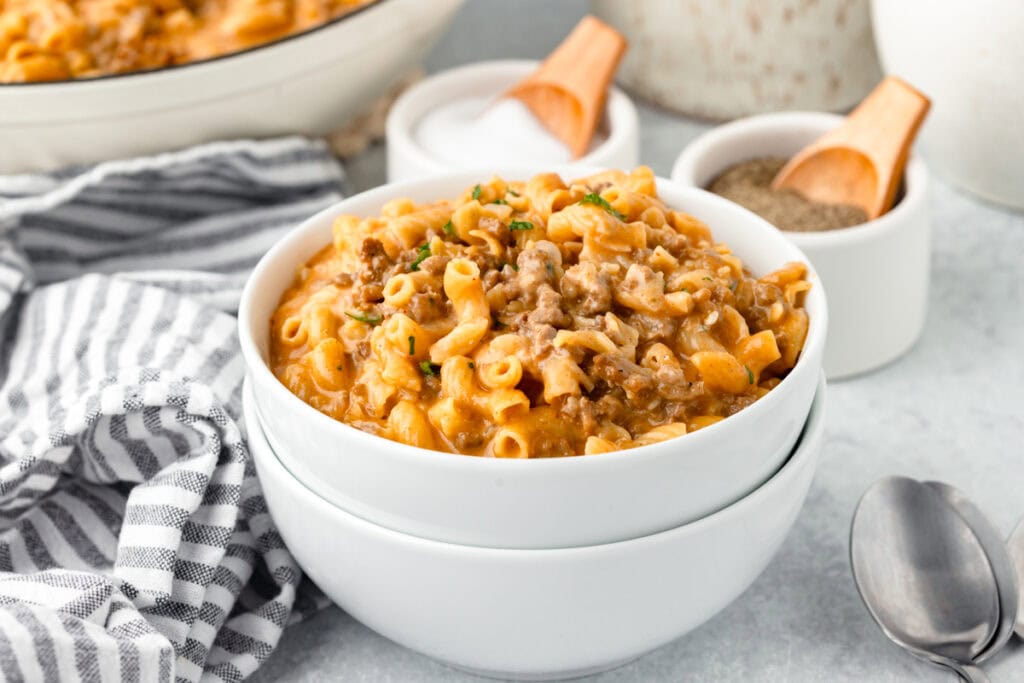 A bowl of creamy beef and macaroni pasta topped with chopped herbs, placed on a table with a striped cloth. Nearby, small bowls contain salt and pepper with wooden scoops. A spoon and fork lay beside them.