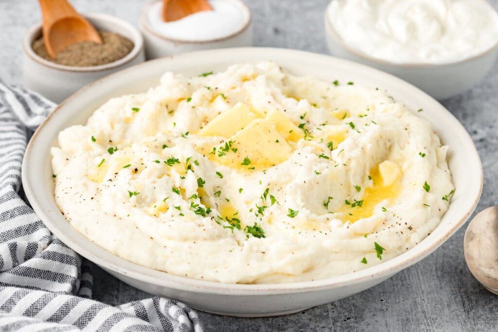 A bowl of creamy mashed potatoes topped with butter and sprinkled with chopped parsley. Salt and pepper in small bowls are in the background next to a dish of sour cream. A striped napkin and a wooden spoon are nearby.