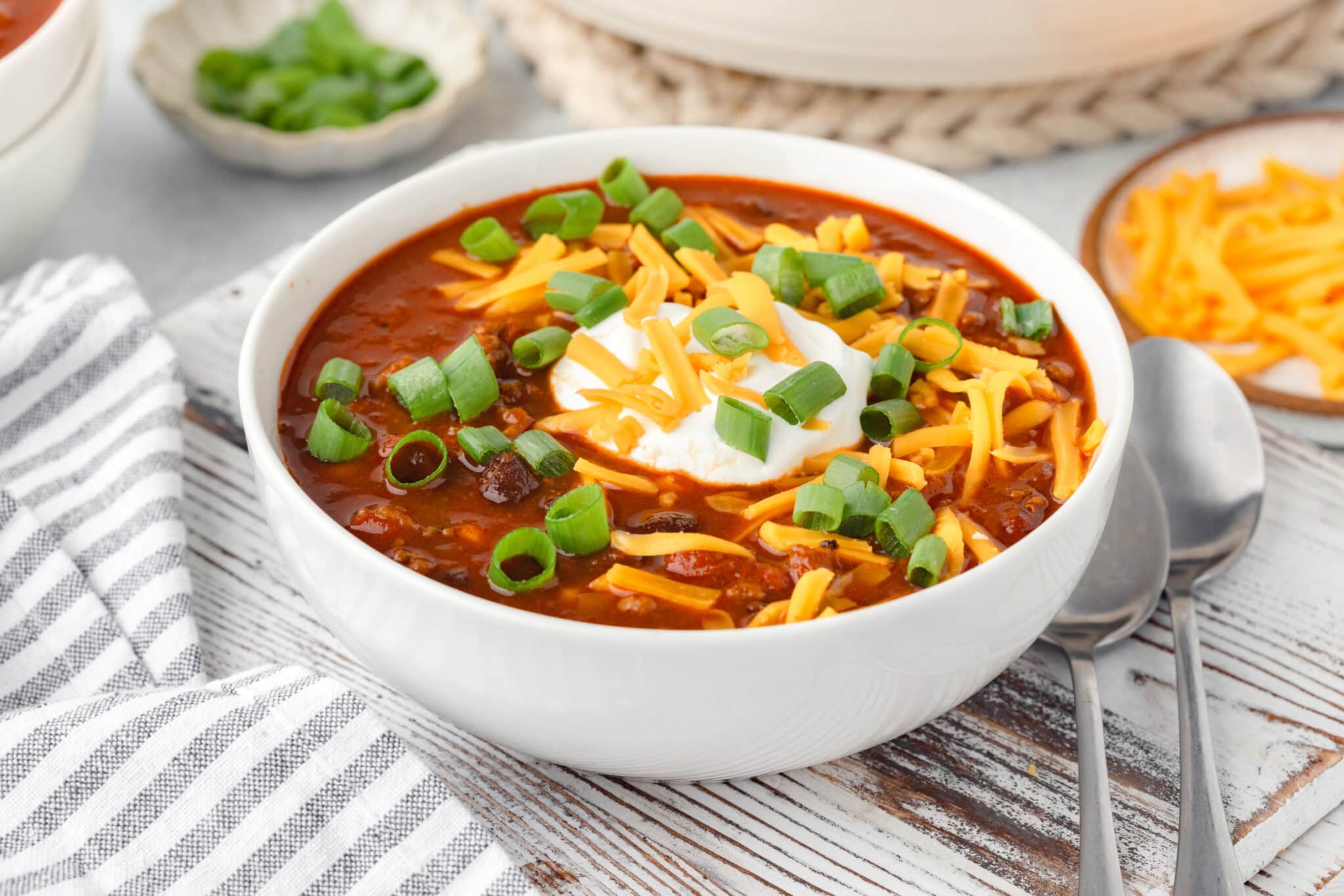 A bowl of chili topped with shredded cheese, sour cream, and chopped green onions sits on a wooden table with a striped napkin and two spoons beside it. In the background, there is a small dish of chopped green onions and the edge of another dish.