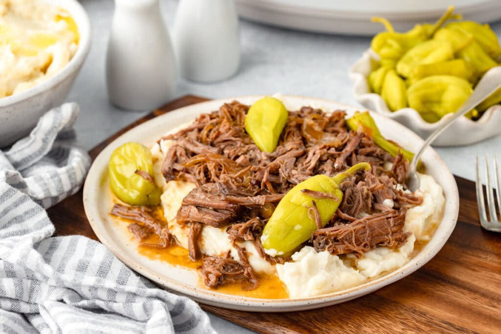 A plate of shredded beef rests on a bed of mashed potatoes, garnished with whole pepperoncini peppers. A fork is placed next to the plate on a wooden cutting board, with a striped napkin nearby. In the background, additional dishes and condiments can be seen.