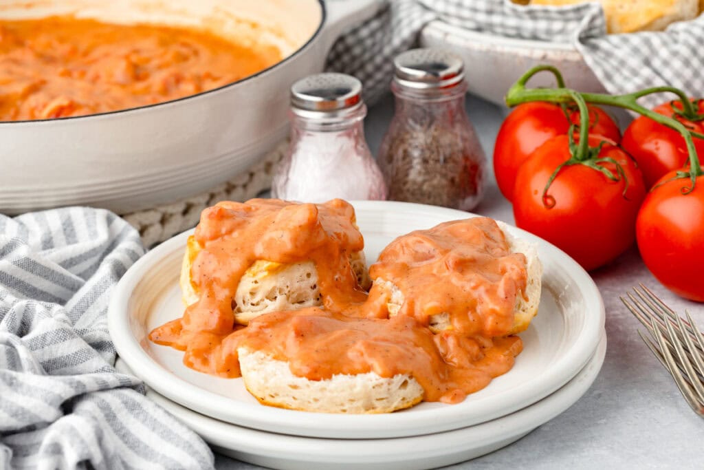 A plate of biscuits topped with creamy red tomato gravy. Fresh tomatoes, a bowl of the sauce, salt and pepper shakers, and a striped cloth are in the background. The scene is set on a light-colored surface.