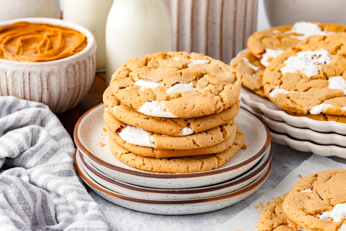 Fluffernutter Cookies stacked on a plate, with a bowl of pumpkin spread and milk bottles in the background.
