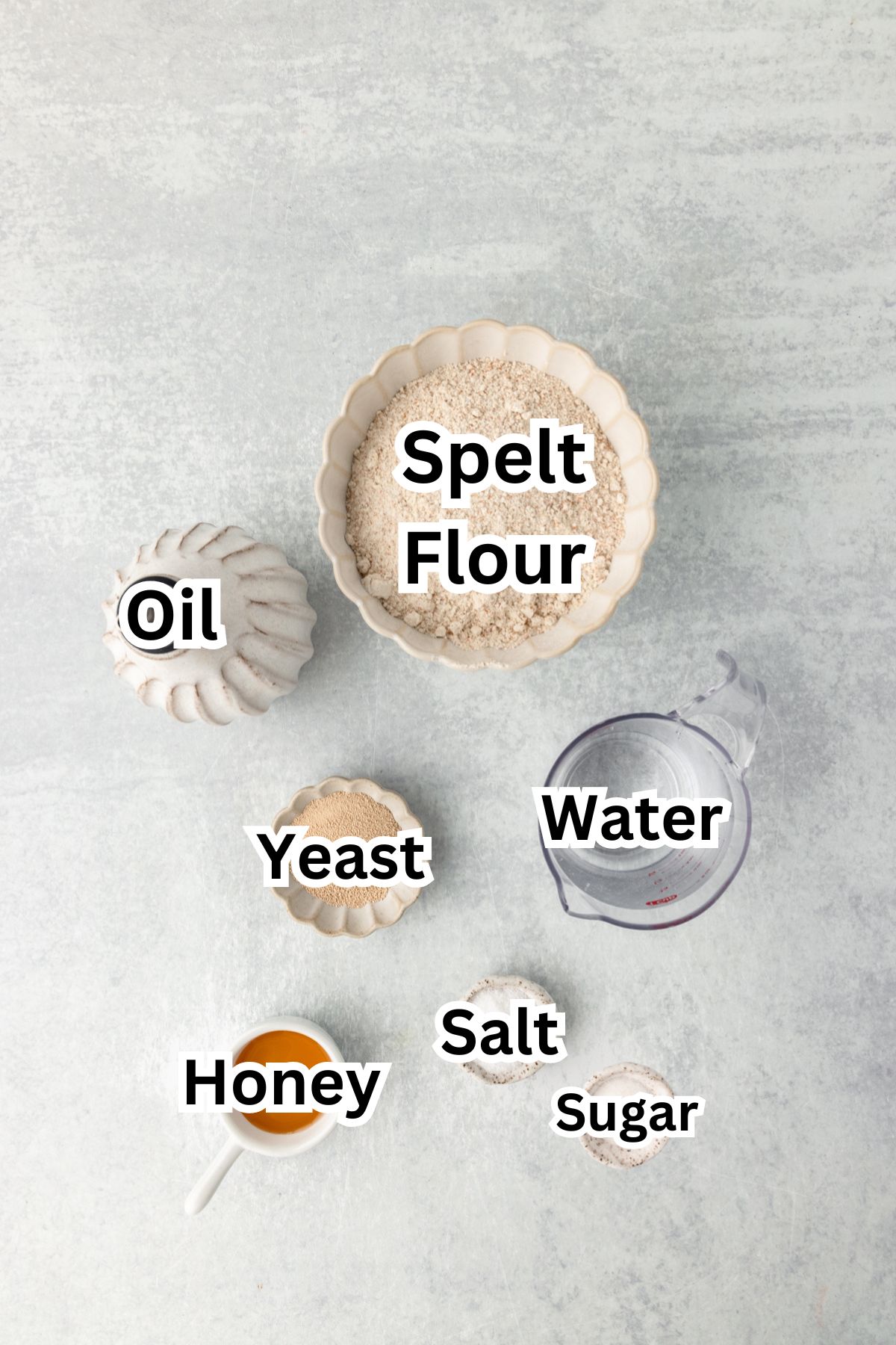 Ground spelt flour with small bowls of honey, yeast, salt, sugar, water, oil, and flour on light gray background.