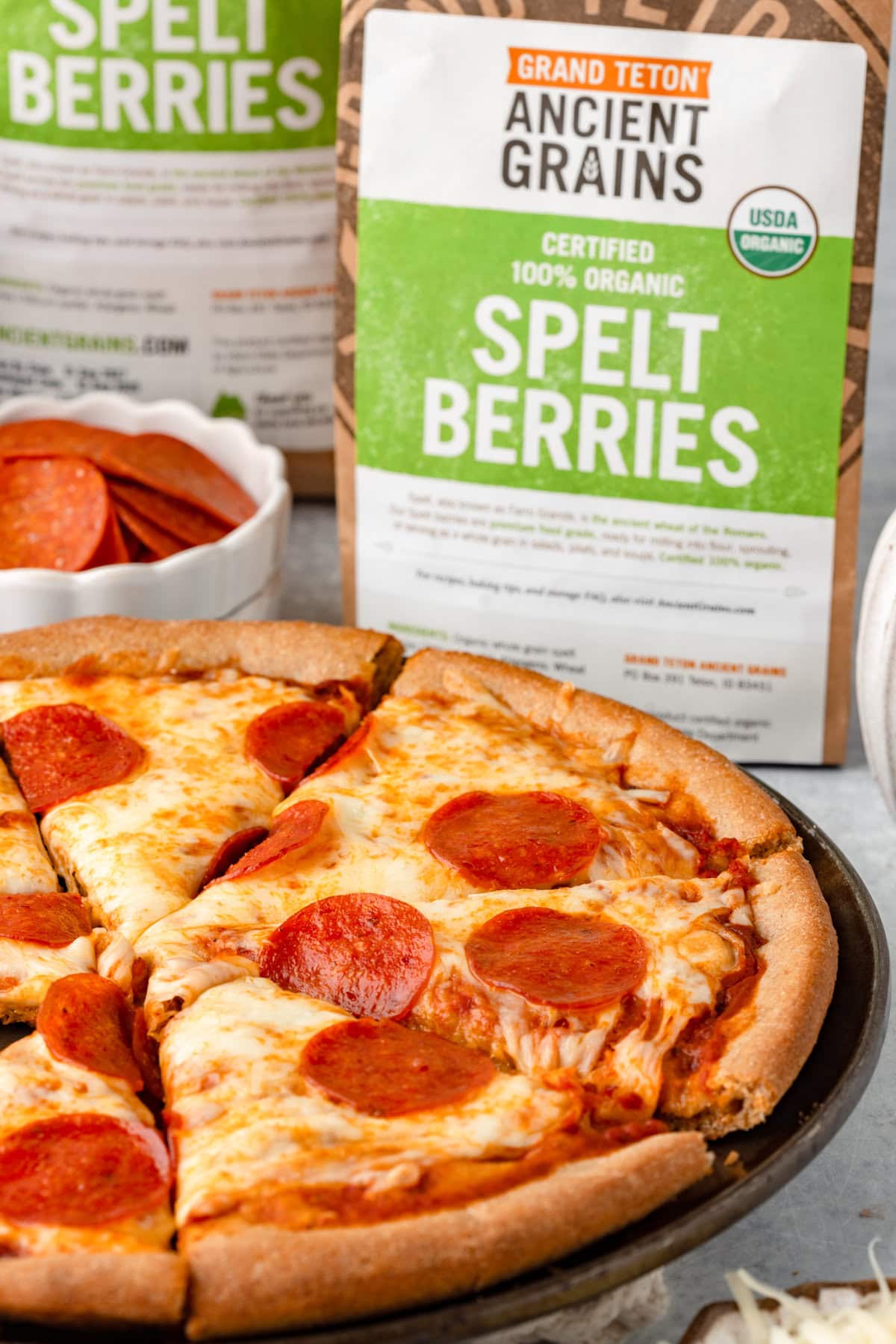 Crunchy gluten-free pepperoni pizza on black dish with organic spelt berries in background.