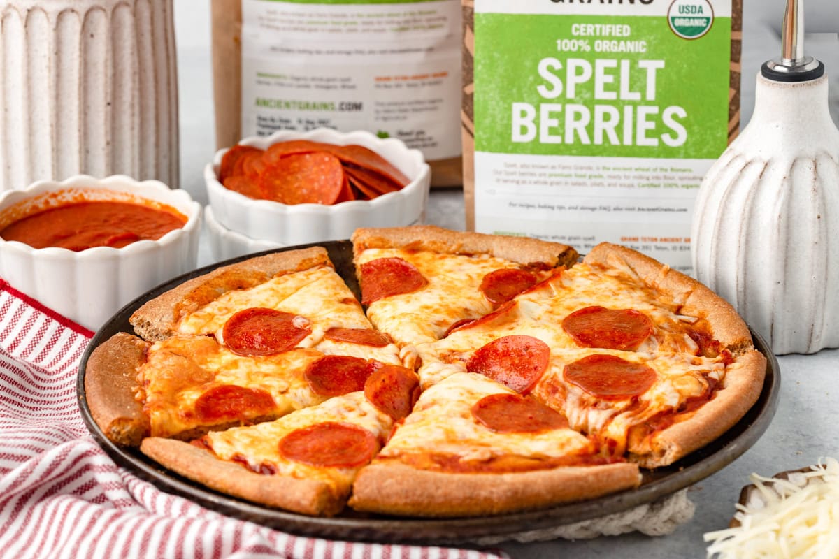 Cheese and pepperoni pizza on a black plate with spelt berries and baking ingredients in background.