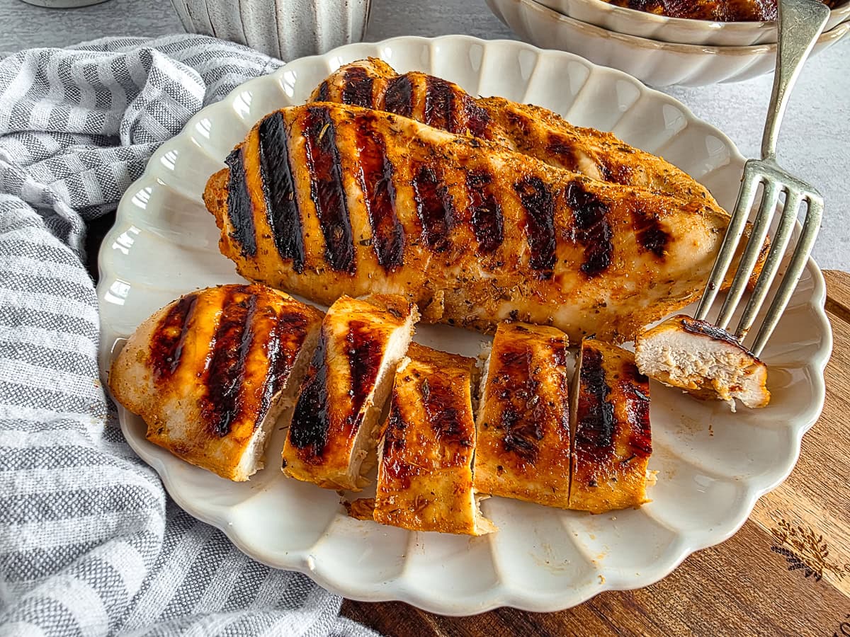 Grilled chicken breast with char marks on a white plate, served with a side dish in the background.
